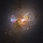 Dwarf starburst galaxy Henize 2-10 seen through the NASA Hubble Space Telescope. The bright region at the center, surrounded by pink clouds and dark dust lanes, indicates the location of the galaxy's massive black hole.