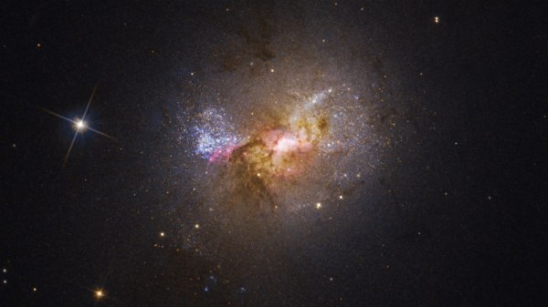 Dwarf starburst galaxy Henize 2-10 seen through the NASA Hubble Space Telescope. The bright region at the center, surrounded by pink clouds and dark dust lanes, indicates the location of the galaxy's massive black hole.