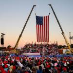A crowd wearing red, white, and blue gathers at a Trump rally with two cranes holding up an American flag as the backdrop.