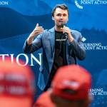 Charlie Kirk speaks with a microphone while people with red MAGA hats watch from the crowd.