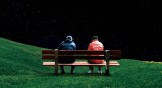 Two people in red and blue jackets sitting on a bench together.