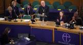 A man in a clergical collar speaks at a podium with the purple Church of England logo.