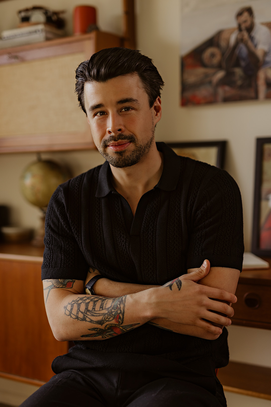 portrait of Josh Nadeau with folded arms against a gallery wall and bookshelf.