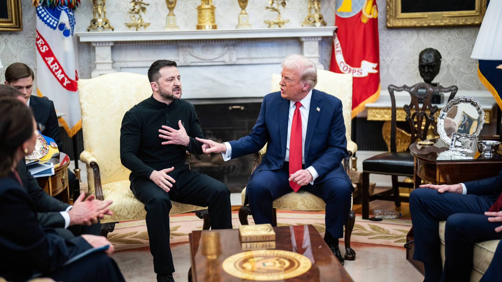 Zelensky and Trump, seated in front of flags, motion to each other while arguing during a meeting.