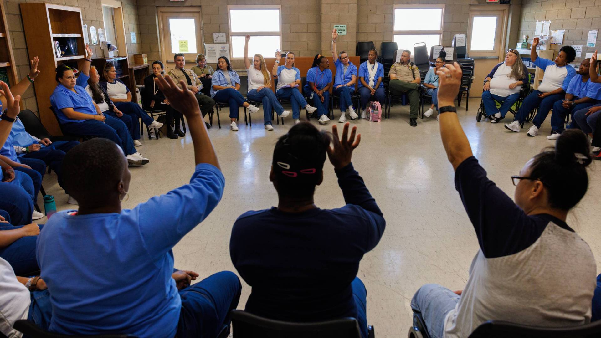 Women in blue uniforms sit in a circle, some raising hands, during a discussion in prison.