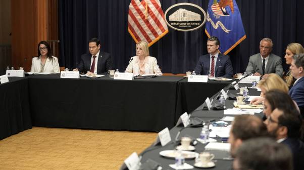 Officials sit in tables in an office with a Justice Department emblem and US flag.