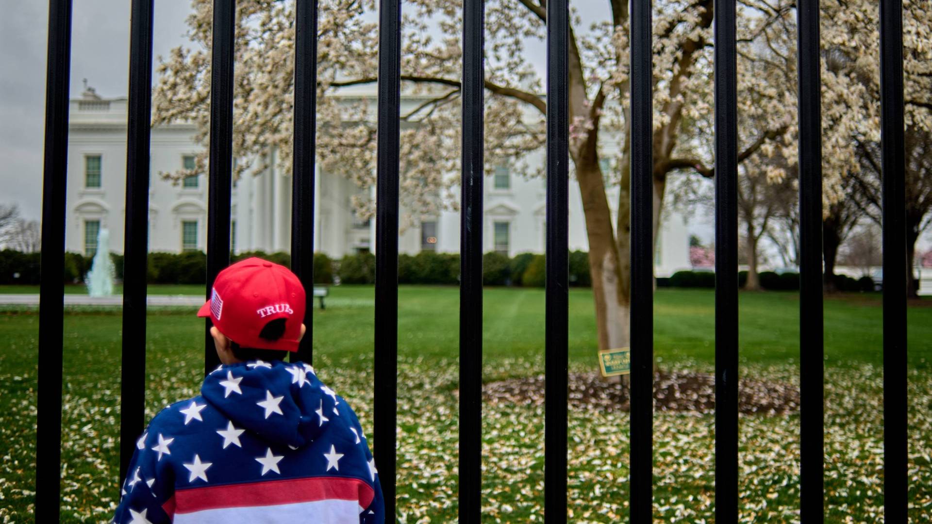 Boy in MAGA hat and flag jacket stands outside White House gate