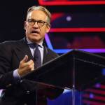 Eric Metaxas speaks, wearing glasses and suit, on a conference stage with neon blue and red background