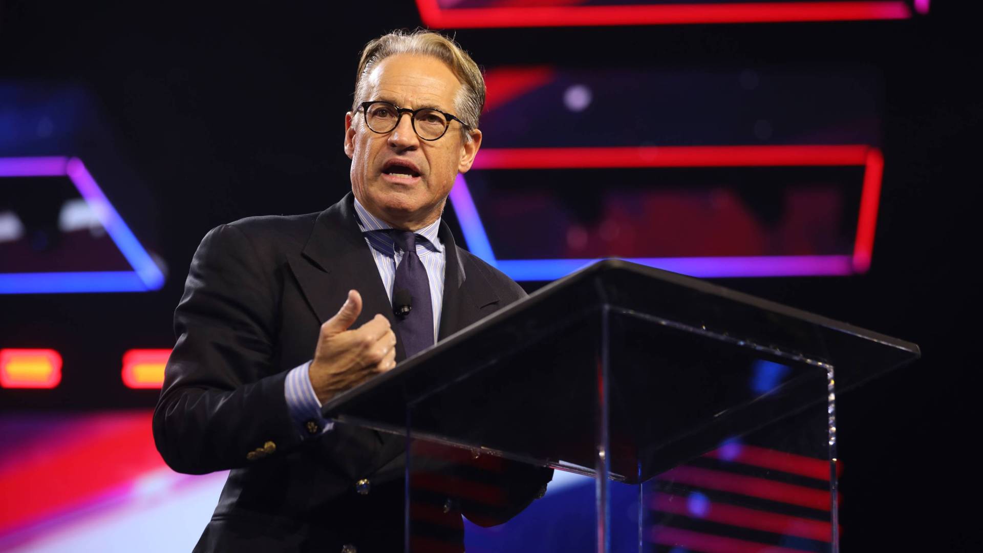 Eric Metaxas speaks, wearing glasses and suit, on a conference stage with neon blue and red background