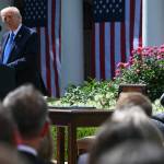 President Trump at podium and Melania Trump seated before a crowd with rose bushes and the White House behind