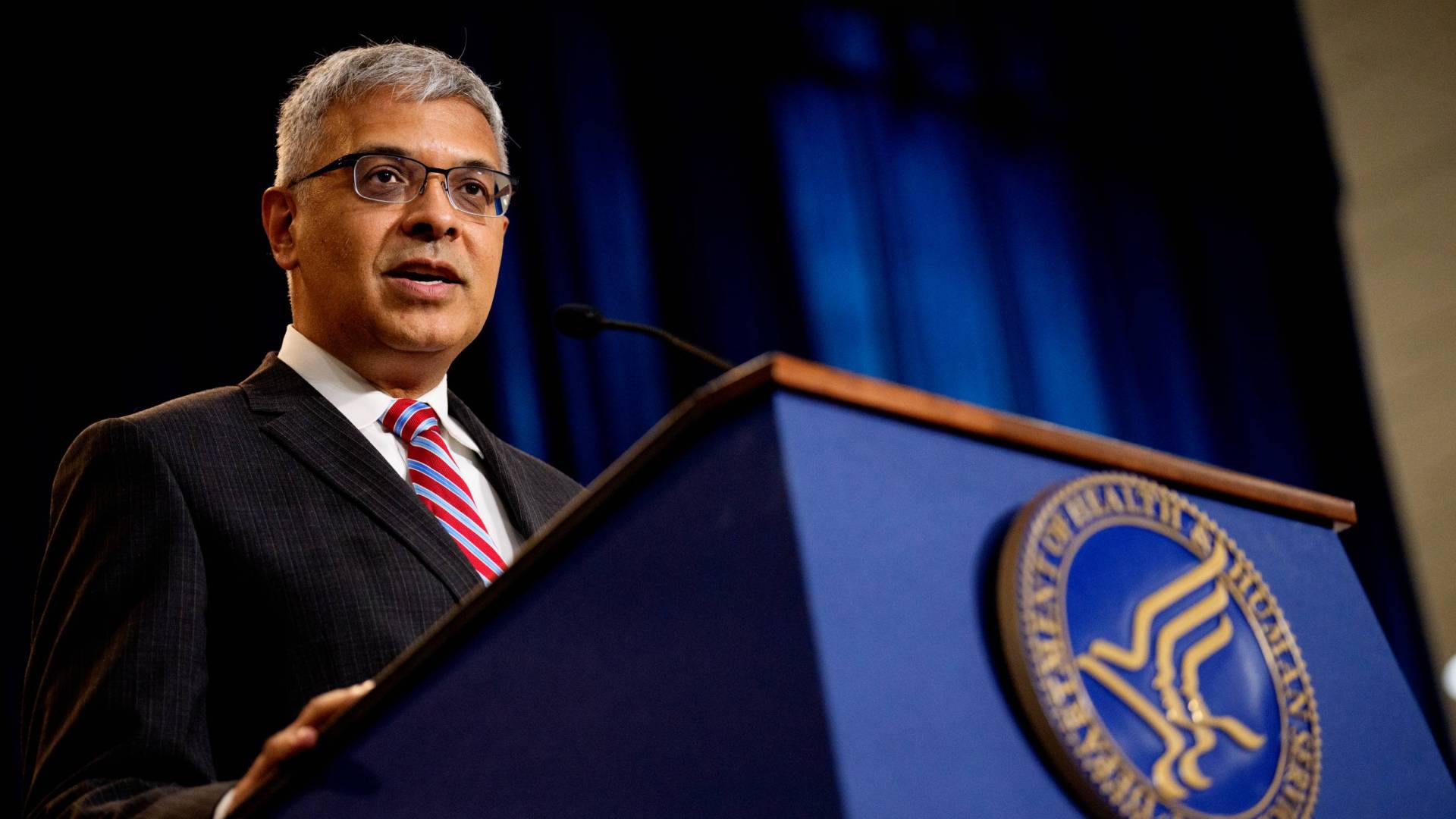 National Institutes of Health director Jay Bhattacharya speaks at a HHS podium with a blue curtain behind.