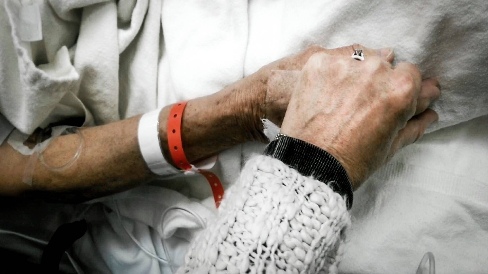 Two people clasp hands on the lap of a hospital patient with a medical bracelet