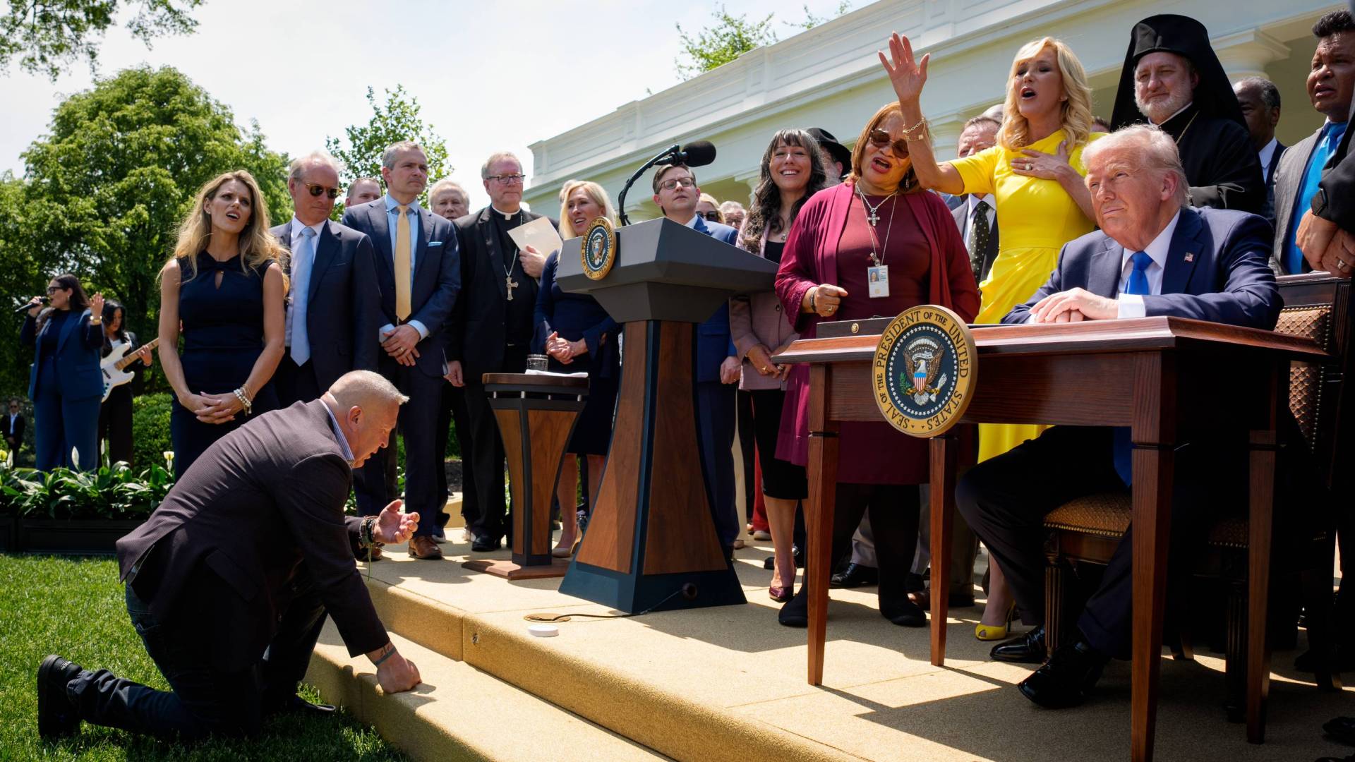 President Trump at a desk in the Rose Garden, surrounded by clergy and ministry leaders, with a man kneeling before him.