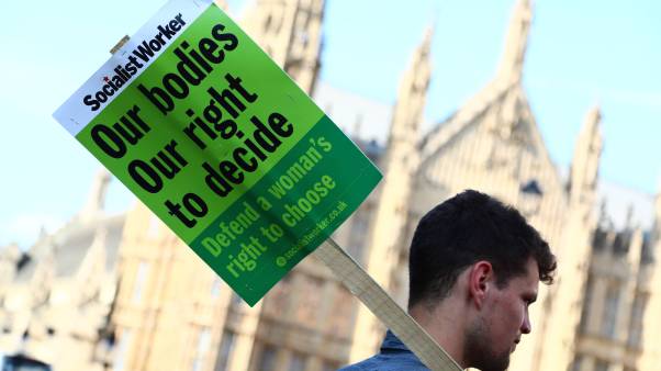 A man holding a green sign reading "our bodies, our right to decide" stands in front of the UK Parliament building.