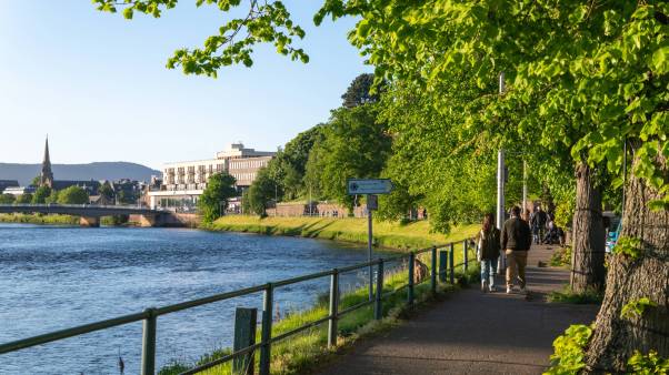 People walk alongside river Ness in Scotland with a church tower in the background.