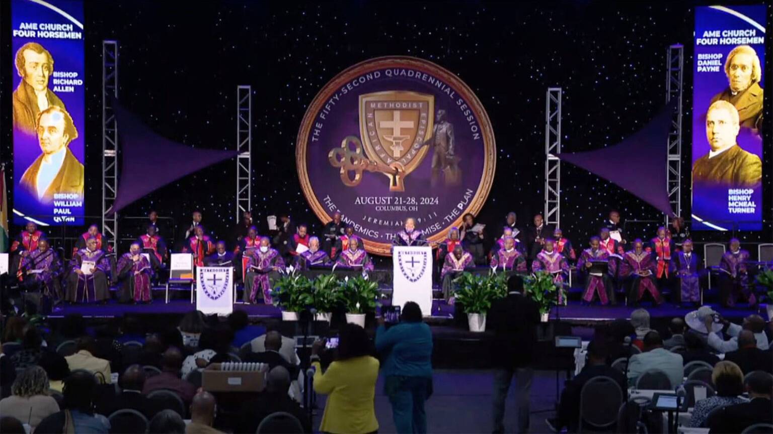 Clergy in purple robes site on a stage behind two clergy at lecturns.