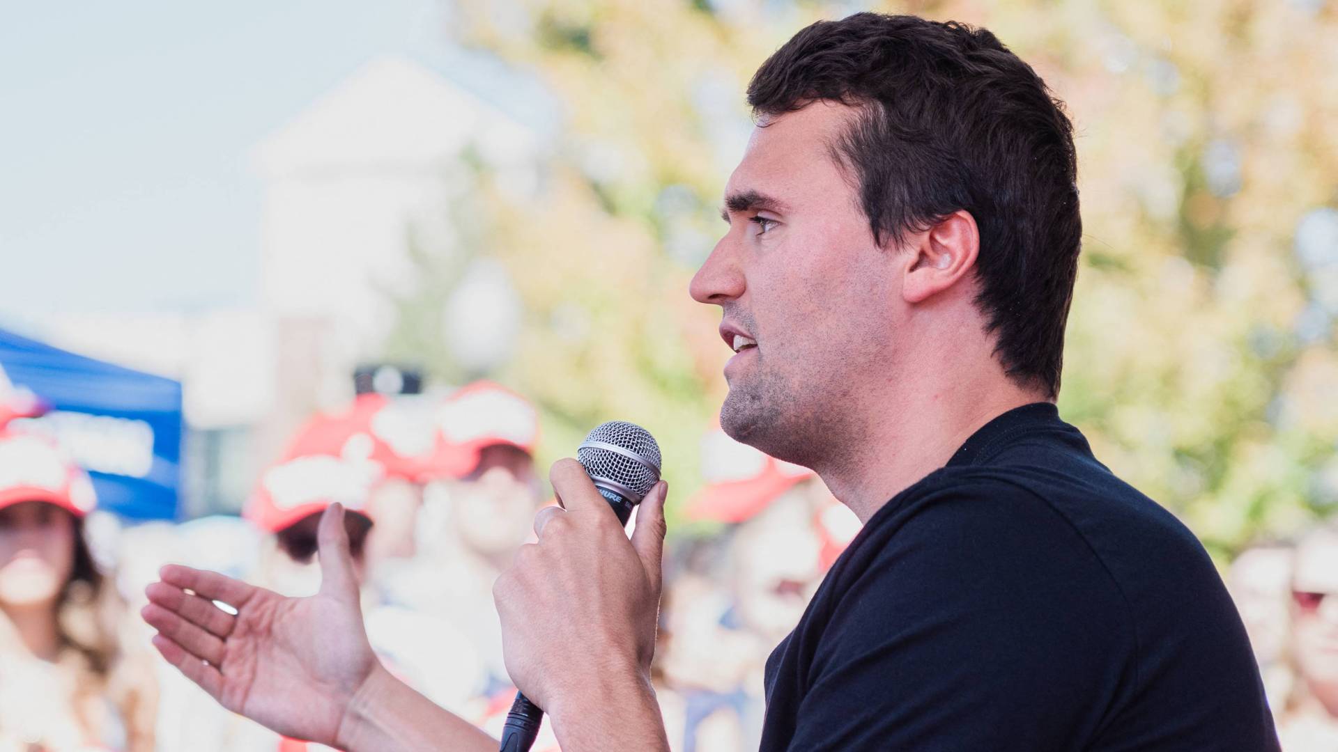 Charlie Kirk in a black T-shirt holding a microphone at an outdoor event.