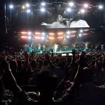 Stage with lights and a crowd with hands raised. Naomi Raine pictured on screen.