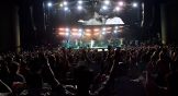 Stage with lights and a crowd with hands raised. Naomi Raine pictured on screen.