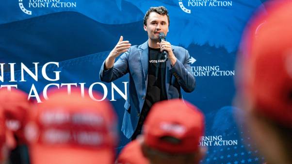 Charlie Kirk holding a microphone in front of a blue Turning Point background with students in red MAGA hats in the foreground.