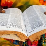 A Bible open in the foreground with hands and colorful dress of a Ugandan woman in the background.
