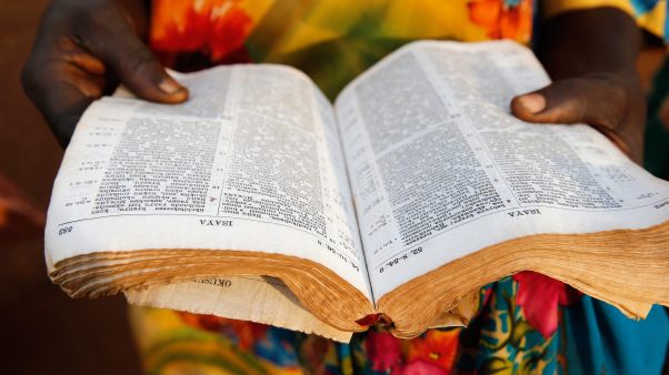 A Bible open in the foreground with hands and colorful dress of a Ugandan woman in the background.