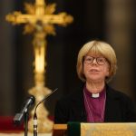 A woman at a lectern in a clerical collar in front of a gold cross.