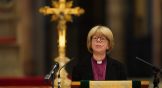 A woman at a lectern in a clerical collar in front of a gold cross.