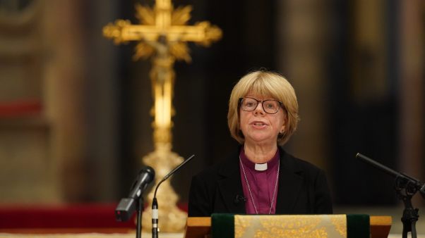 A woman at a lectern in a clerical collar in front of a gold cross.