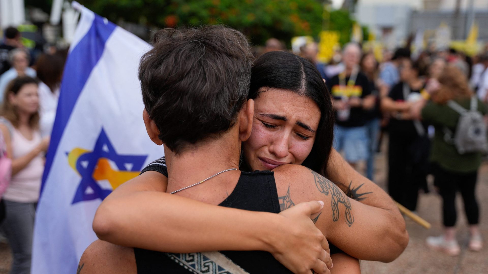 A tearful woman hugs a man in Tel Aviv while responding to news of a ceasefire deal. Israeli flag behind them.