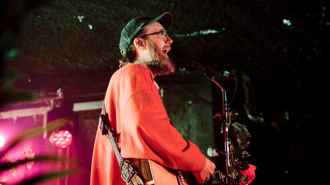 Side profile of John Mark McMillan playing guitar on stage in a red jacket and black cap.