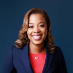 Nicole Massie Martin smiles wearing a red shirt and blue blazer against a blue background.
