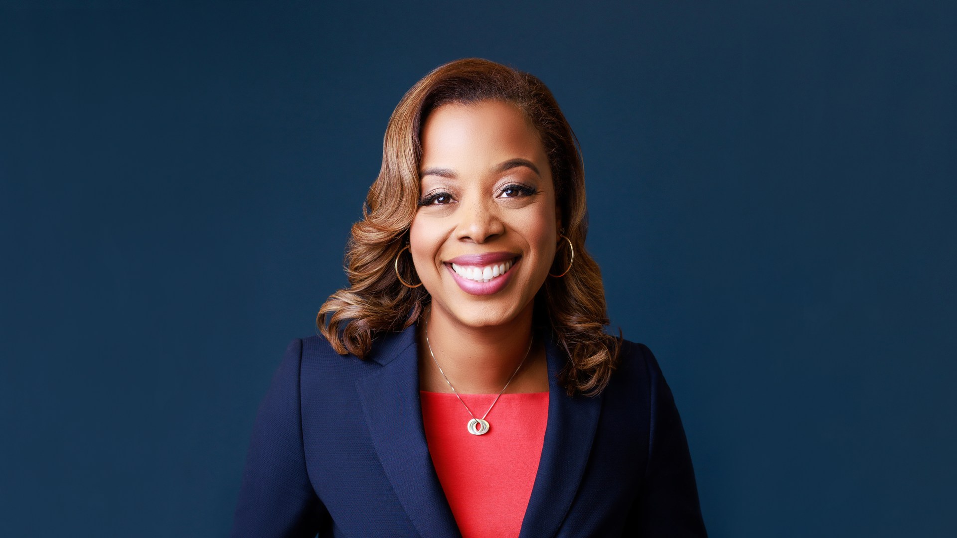 Nicole Massie Martin smiles wearing a red shirt and blue blazer against a blue background.