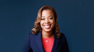 Nicole Massie Martin smiles wearing a red shirt and blue blazer against a blue background.