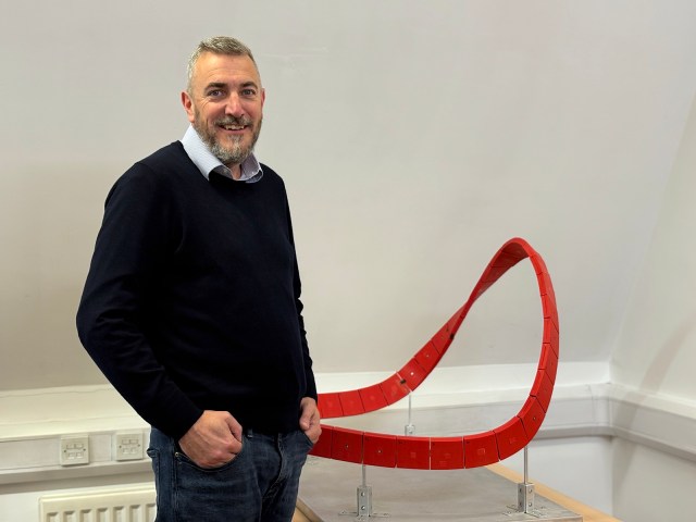 A bearded man in his 50s stands besides a red model shaped like a Möbius strip.