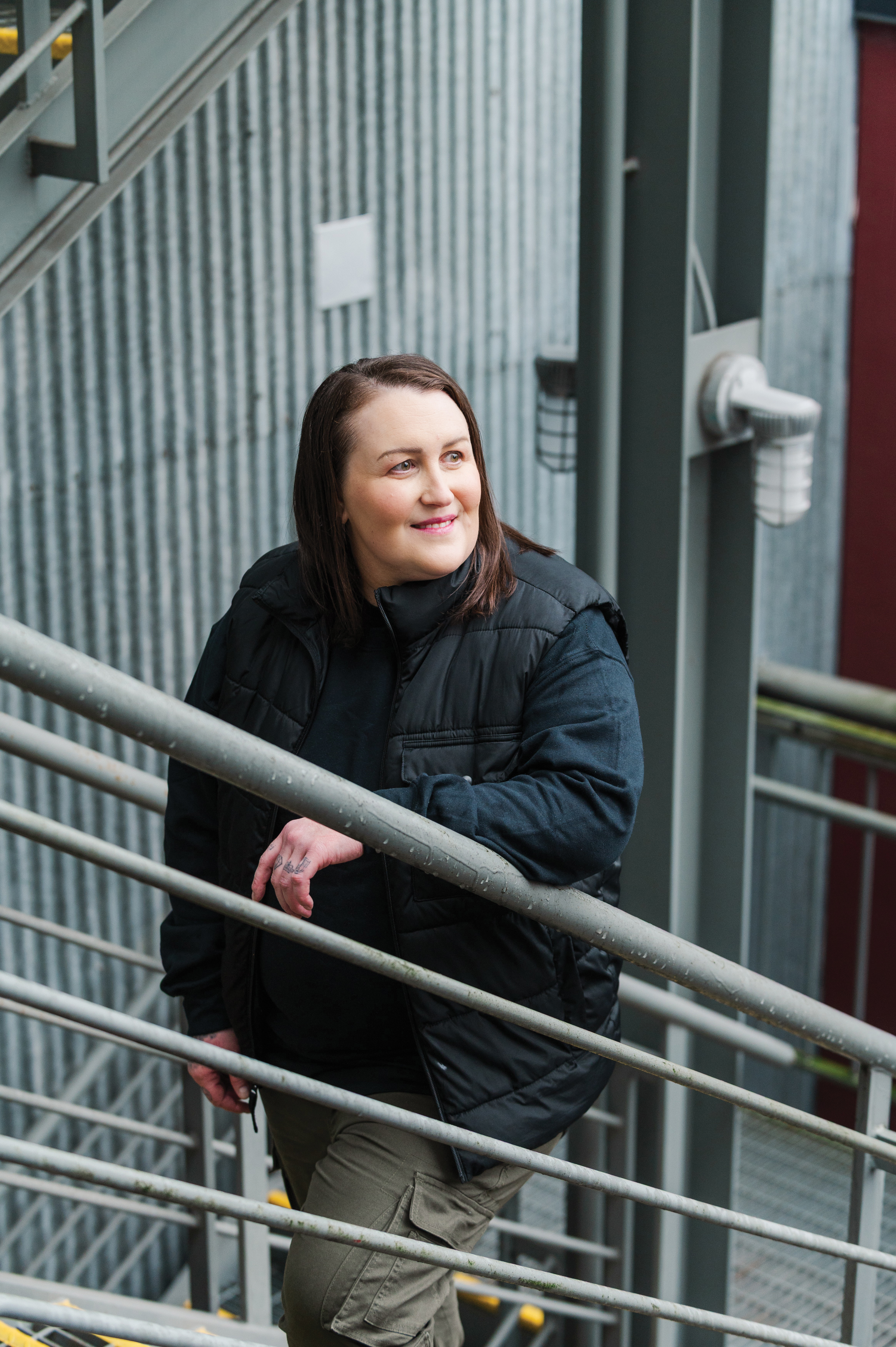Kyla Gillespie in a black jacket standing on a metal staircase in an industrial setting, looking to the side.