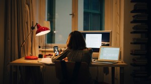 A girl on her phone sitting at a desk with two computers open.