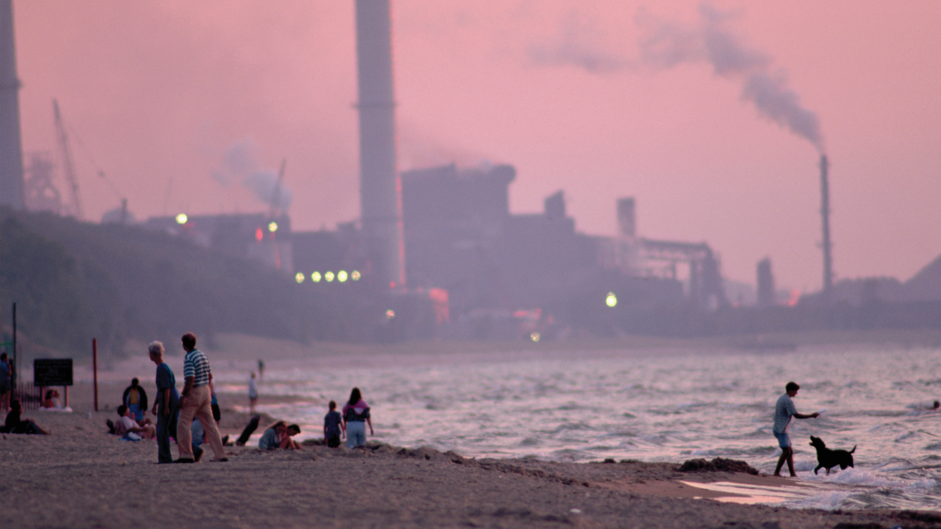 People walk along a sandy beach at dusk while a smokestack-laden industrial plant looms in the background under a pink, hazy sky in Gary, Indiana.